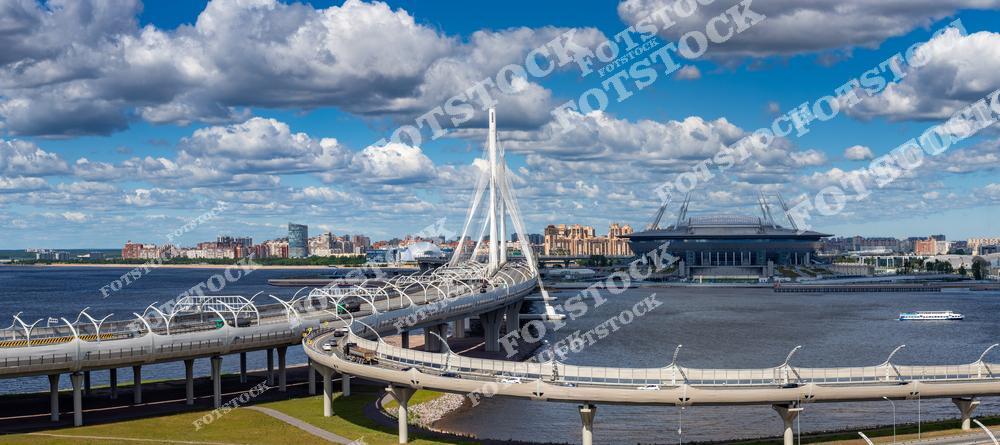 На фотографии изображен Вантовый мост, также известный как Большой Обуховский мост, соединяющий берега Санкт-Петербурга. На заднем плане виден стадион, расположенный на Крестовском острове, а также панорама Финского залива. Яркий солнечный день подчеркивает красоту архитектуры и природы города. Элементы современной инфраструктуры гармонично вписываются в городской пейзаж.